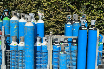 Plusieurs bouteilles de gaz industrielles bleues regroupées dans des racks métalliques en extérieur, devant une haie verte. Certaines bouteilles sont grandes, d’autres plus petites, toutes équipées de détendeurs ou de bouchons protecteurs