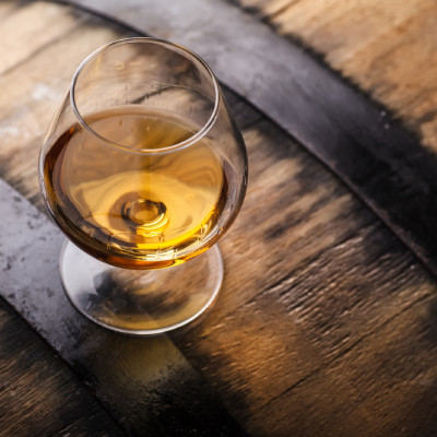 Image montrant un verre de cognac ou de whisky posé sur une surface en bois, probablement un tonneau. Le verre est de forme arrondie, typique pour les spiritueux, et contient un liquide ambré qui capte la lumière de manière élégante. Le fond en bois sombr