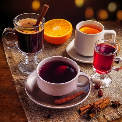 Image montrant plusieurs boissons chaudes servies dans différentes tasses et verres. On y voit du vin chaud dans un verre transparent avec un bâton de cannelle, une tasse de thé ou de tisane avec une tranche d'orange flottant à la surface, ainsi qu'une pe