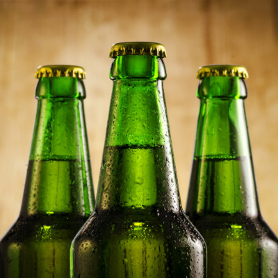 Photo montrant trois bouteilles de bière en verre vert, fermées par des capsules dorées, et recouvertes de fines gouttelettes d'eau. L'arrière-plan est flou, avec des teintes chaudes, contrastant avec l'aspect rafraîchissant des bouteilles. La composition
