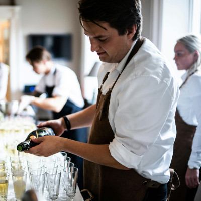 Image montrant un serveur versant du vin ou du champagne dans des verres alignés sur une table. Le serveur porte un tablier marron par-dessus une chemise blanche, suggérant un cadre professionnel, probablement lors d'un événement ou d'une réception. À l'a