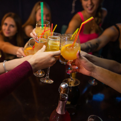 Photo montrant un groupe de personnes trinquant avec des cocktails et du champagne lors d'un enterrement de vie de jeune fille. Les verres colorés et festifs, avec des pailles, ajoutent une ambiance conviviale. Les participantes, souriantes et visiblement