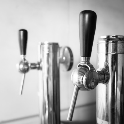 Photo en noir et blanc montrant deux tireuses à bière chromées, avec des poignées noires élégantes. Le premier plan met en avant un des robinets de distribution, bien net et détaillé, tandis que le second robinet apparaît légèrement flou en arrière-plan, 