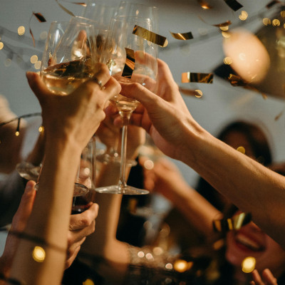 Photo montrant un groupe de personnes levant leurs verres pour un toast, créant une ambiance festive. Les mains tiennent des verres à pied remplis de vin blanc, et des confettis dorés flottent dans l'air, ajoutant une touche de célébration. L'éclairage do